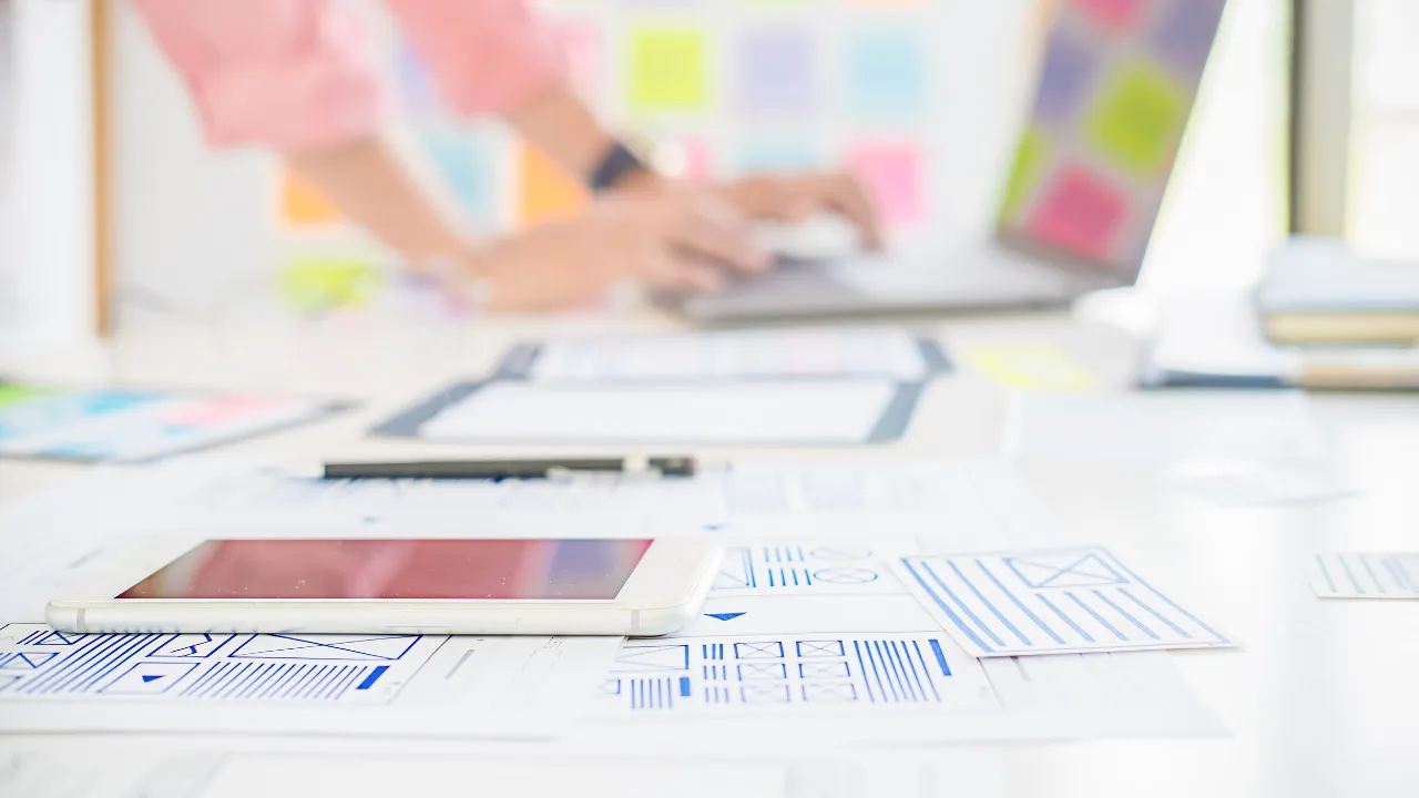 Vue d'un bureau de designer avec des maquettes de sites web dessinées sur des feuilles, une main travaillant sur un ordinateur portable, et des post-its de recherche UX.